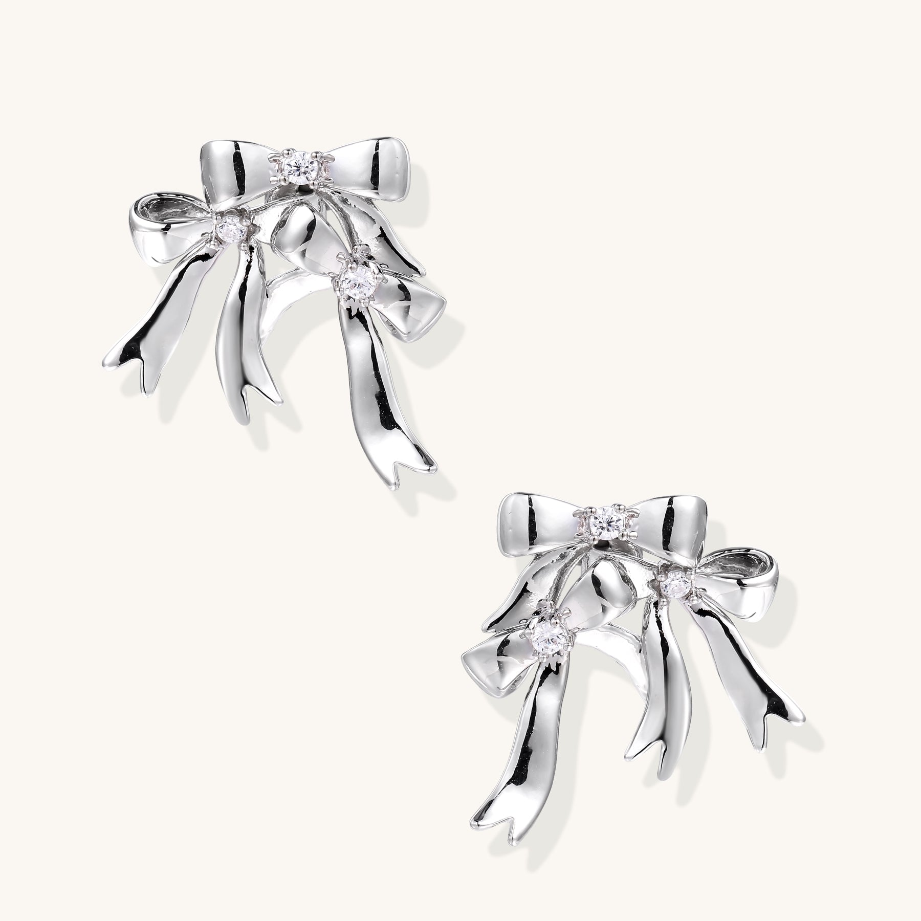 Pair of 18K gold plated brass bow-shaped earrings with flowing ribbon tails and zirconia crystal accents on white background.