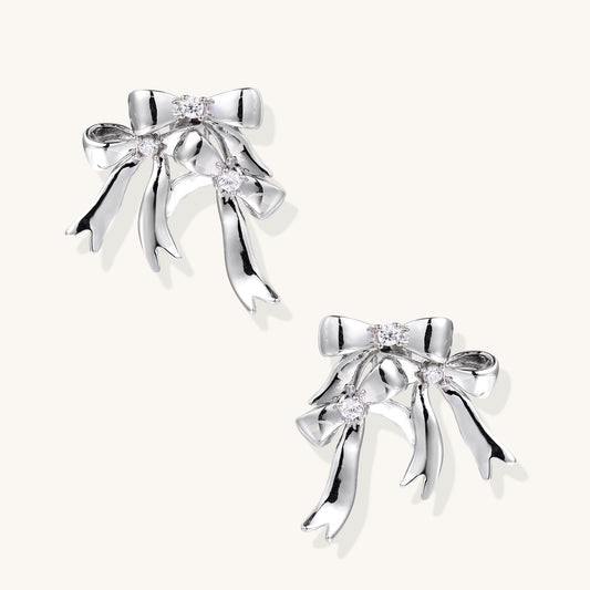 Pair of 18K gold plated brass bow-shaped earrings with flowing ribbon tails and zirconia crystal accents on white background.